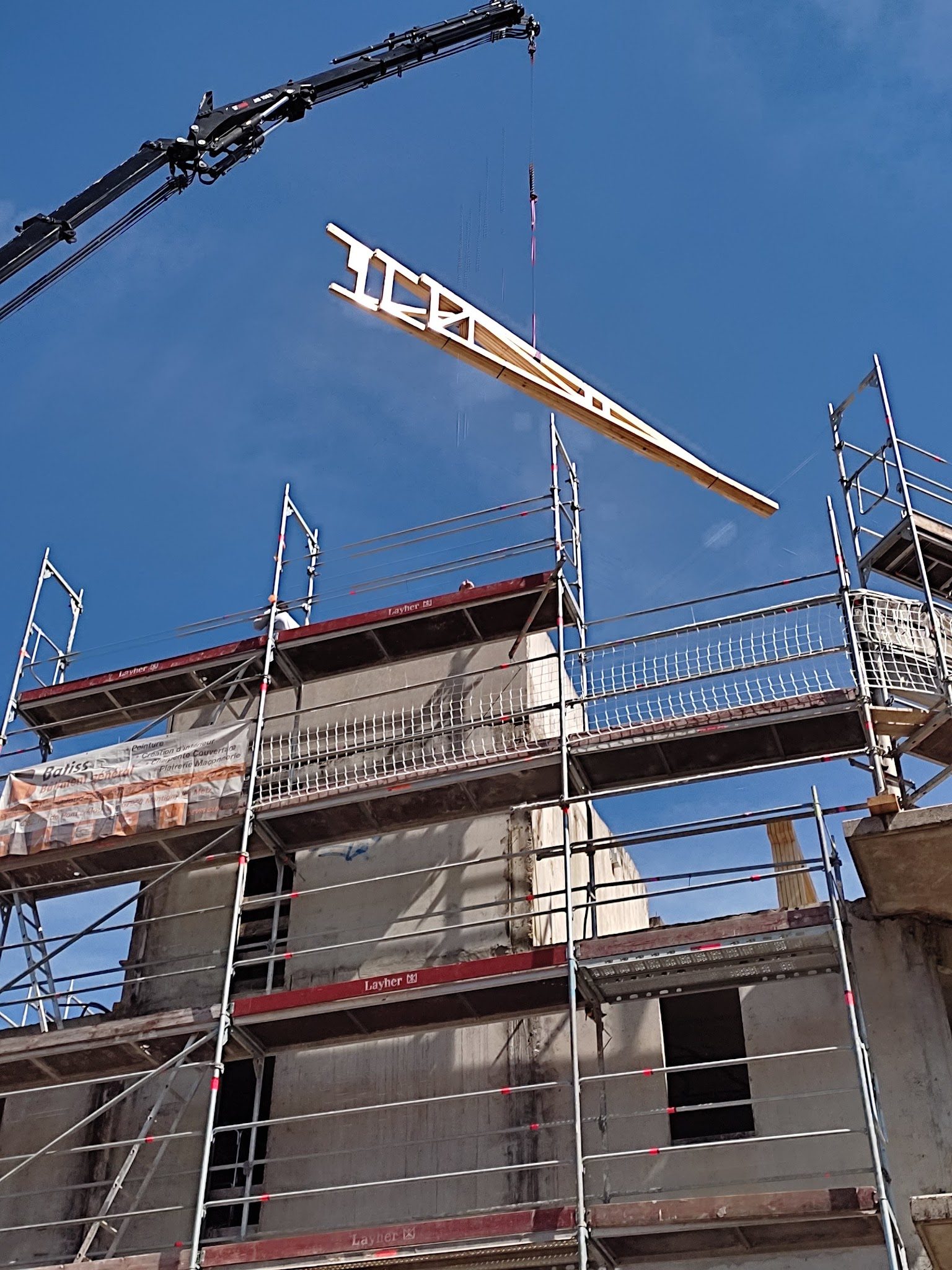 Ferme de charpente levée par un bras de grue au-dessus d'un immeuble sous échafaudage